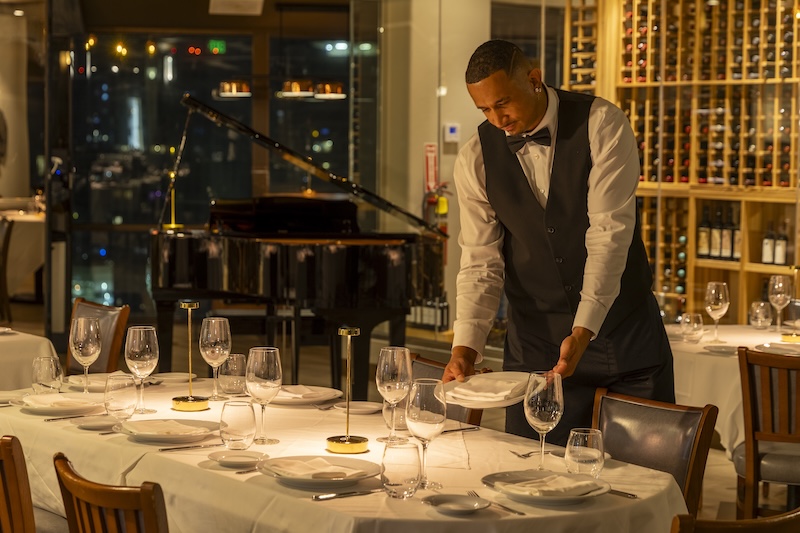 Server setting a table at Empire Steak House Hawaii with Honolulu night views behind